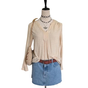 May include: A beige long-sleeve top with a V-neck, paired with a denim mini-skirt and a brown belt. A small, woven shoulder bag and layered necklaces complete the outfit. The mannequin is on a white background.