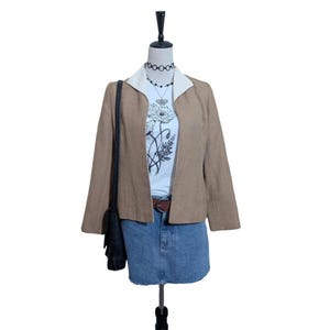 May include: A tan blazer with a white collar is worn over a white t-shirt with a floral design. A denim skirt and a black shoulder bag complete the outfit. A necklace and belt are also visible.