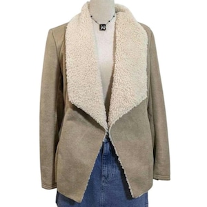 May include: A tan suede-like jacket with a cream-colored, fluffy shearling collar. The open-front jacket is displayed on a mannequin, revealing a glimpse of a denim skirt. A black necklace with a square pendant is visible.