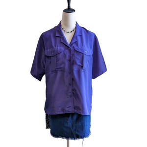 May include: A short-sleeved, purple button-down shirt with two chest pockets. The shirt is displayed on a mannequin, paired with a denim skirt. A black beaded necklace is visible.
