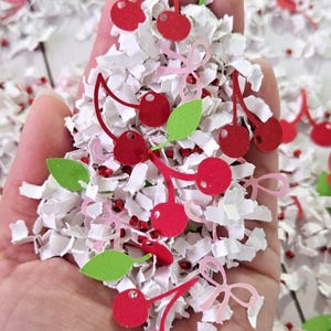 Op de afbeelding: Een hand die een stapel rode en groene papieren kersconfetti vasthoudt. De confetti ligt op een wit oppervlak.