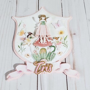 以下が含まれることがあります： 妖精をテーマにした、盾型のレイヤーケーキトッパー。妖精、花、キノコが特徴です。「Tris」という名前のピンクとゴールドのバナーが含まれています。カラーパレットはソフトで、ピンク、グリーン、イエローを使用しています。