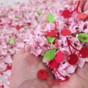 Op de afbeelding: Een hand die roze en groene confetti vasthoudt met rode kersenvormen. De confetti is gemaakt van papier en is perfect om elke gelegenheid een feestelijke touch te geven.
