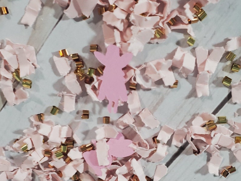 May include: Pink and gold confetti with a pink paper cutout of a fairy. The fairy has wings and a dress.