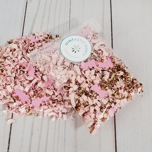 May include: A clear plastic bag filled with pink and gold confetti. The confetti is made of paper and includes small gold beads and pink paper cutouts. The bag has a sticker on it that says "Mintastic Party".