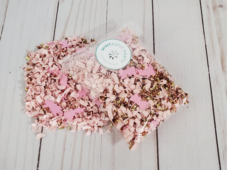 May include: A clear plastic bag filled with pink and gold confetti. The confetti is made of paper and includes small gold beads and pink paper cutouts. The bag has a sticker on it that says "Mintastic Party".