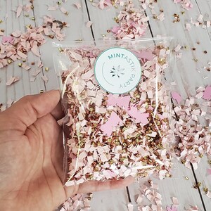 May include: A clear plastic bag filled with pink and gold confetti. The confetti is made of paper and has a butterfly shape. The bag has a sticker on it that says "Mintastic Party".
