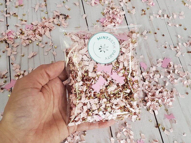 May include: A clear plastic bag filled with pink and gold confetti. The confetti is made of paper and has a butterfly shape. The bag has a sticker on it that says "Mintastic Party".
