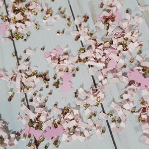 May include: Pink and gold confetti with a variety of shapes, including small strips and larger cut-out shapes. The confetti is scattered on a white wooden surface.