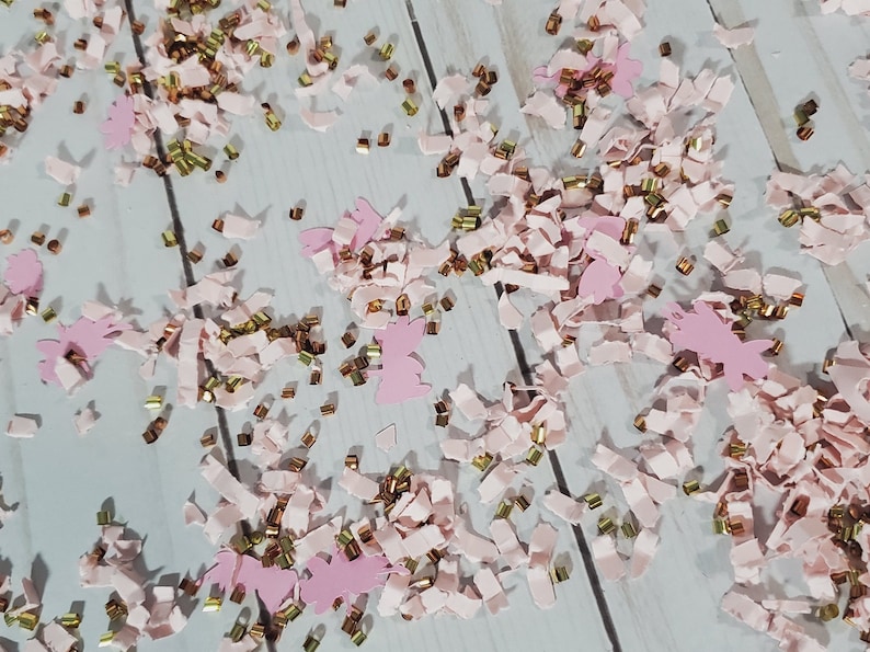 May include: Pink and gold confetti with a variety of shapes, including small strips and larger cut-out shapes. The confetti is scattered on a white wooden surface.