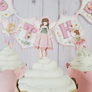 May include: Three pink and white cupcakes with fairy themed toppers. The center cupcake has a pink fairy with brown hair and green wings. The cupcakes are decorated with white frosting and glitter.