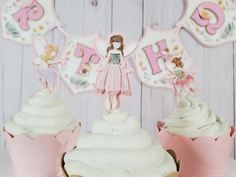 May include: Three pink and white cupcakes with fairy themed toppers. The center cupcake has a pink fairy with brown hair and green wings. The cupcakes are decorated with white frosting and glitter.