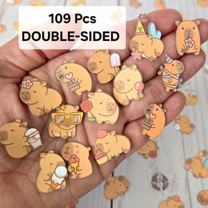 May include: A collection of 109 double-sided capybara stickers in various poses, some holding treats or wearing accessories. The stickers are primarily tan and orange with black outlines, and the text "109 Pcs DOUBLE-SIDED" is visible.