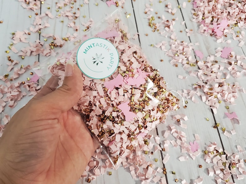 May include: A clear plastic bag filled with pink and gold confetti. The confetti is made of paper and includes small pieces of paper, stars, and butterflies. The bag has a sticker on it that says "Mintastik Party".