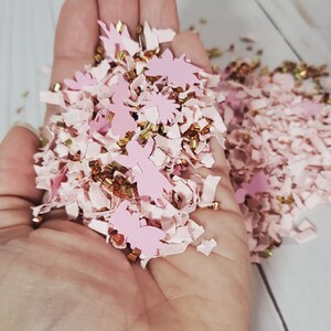 May include: Pink and gold confetti with a variety of shapes, including small stars, hearts, and flowers. The confetti is in a hand, ready to be scattered.