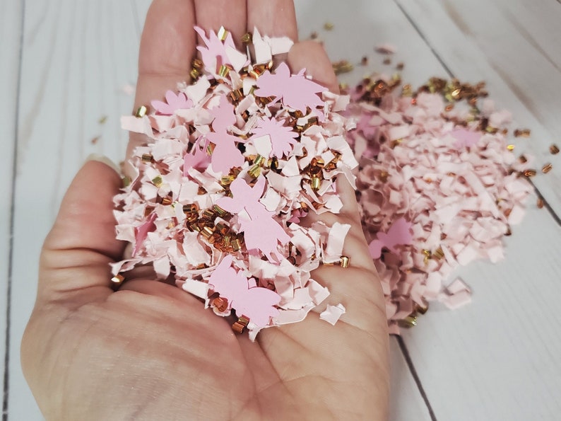May include: Pink and gold confetti with a variety of shapes, including small stars, hearts, and flowers. The confetti is in a hand, ready to be scattered.