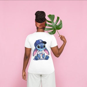 Op de afbeelding: Wit T-shirt met een blauw Stitch-personage dat een baseballpet en een shirt van de Los Angeles Dodgers draagt. Het personage zit met gekruiste benen.