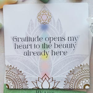 May include: A white card with the text "Gratitude opens my heart to the beauty already here." The card features a lotus flower design with gold and pastel accents. The word "HEART" is at the bottom.
