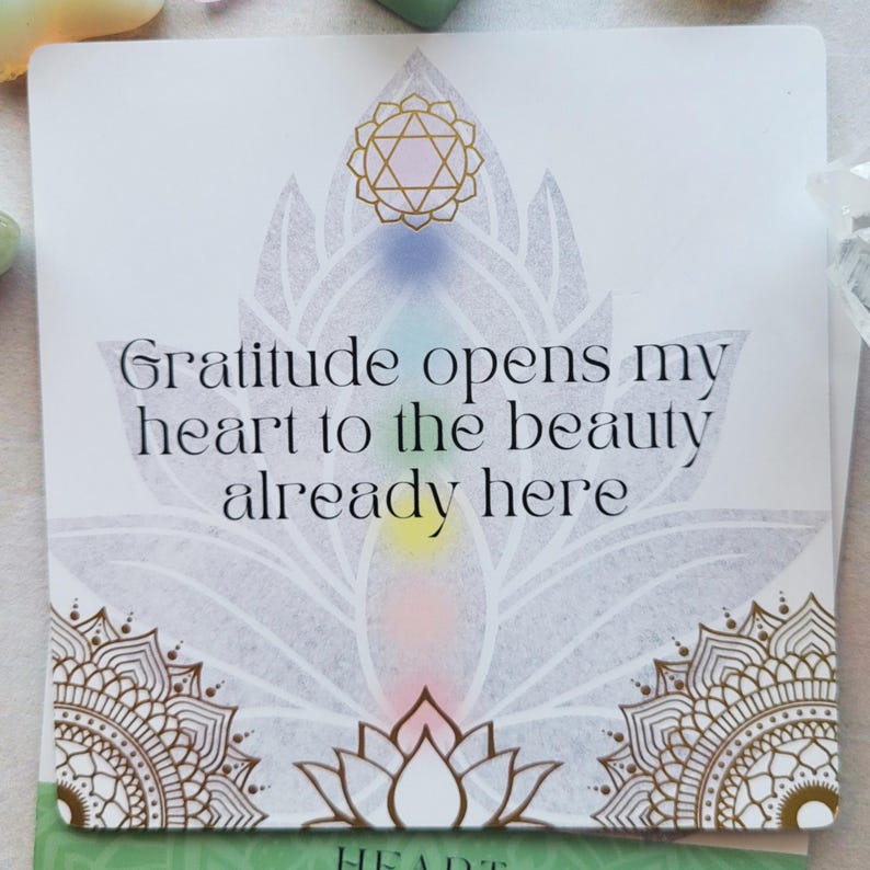 May include: A white card with the text "Gratitude opens my heart to the beauty already here." The card features a lotus flower design with gold and pastel accents. The word "HEART" is at the bottom.