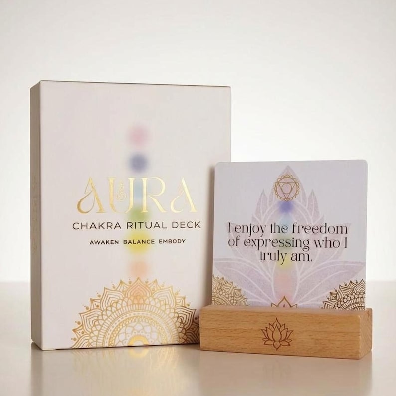May include: A white Aura Chakra Ritual Deck box with gold lettering and a decorative mandala design. A card with the text "I enjoy the freedom of expressing who I truly am" is displayed on a wooden stand. The deck is designed to Awaken, Balance, and Embody.