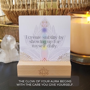 May include: A white card with the text "I create stability by showing up for myself daily" on a wooden stand. The card features a lotus flower design with pastel colors and a geometric symbol. The bottom of the card reads "THE GLOW OF YOUR AURA BEGINS WITH THE CARE YOU GIVE YOURSELF."