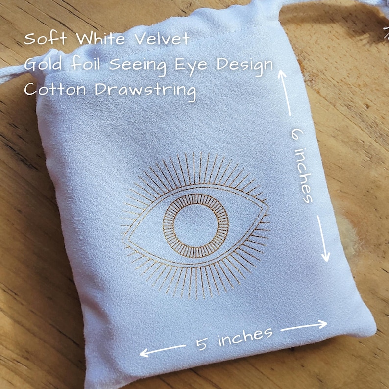 May include: A white velvet drawstring bag with a gold foil seeing eye design. The bag is 5 inches wide and 6 inches tall. The text "Soft White Velvet", "Gold foil Seeing Eye Design", and "Cotton Drawstring" are printed on the bag.