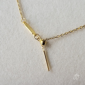 May include: Close-up of a delicate gold necklace with a small chain and a vertical bar pendant. The necklace features a sliding bead and a small, dangling bar. The jewelry is set against a neutral background.