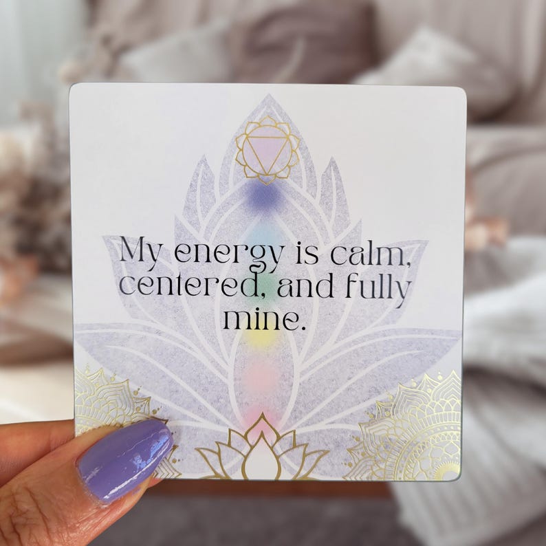 May include: A square card with a lotus flower design and the text "My energy is calm, centered, and fully mine." The card features a watercolor effect with pastel colors and gold accents. The card is held by a person with purple nail polish.