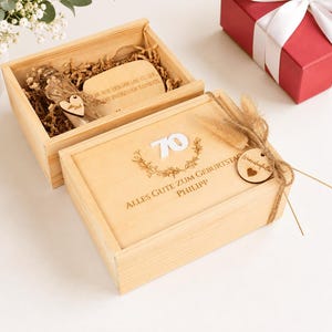 Könnte beinhalten: Eine helle Holzgeschenkbox mit Schiebedeckel, graviert mit der Zahl "70", dem Text "ALLES GUTE ZUM GEBURTSTAG" und "PHILIPP" sowie einem Blumenkranz. Ein kleines Holzschild mit der Aufschrift "Wunscherfüller" ist mit Kordel befestigt.