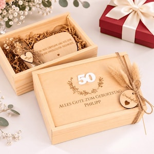 Könnte beinhalten: Eine helle Holzgeschenkbox mit Schiebedeckel, graviert mit der Zahl 50, einem Blumenkranz und dem Text ALLES GUTE ZUM GEBURTSTAG PHILIPP. Die Box ist mit einer Schnur und einem Holzanhänger versehen und enthält eine Glasflasche sowie eine Holztafel.