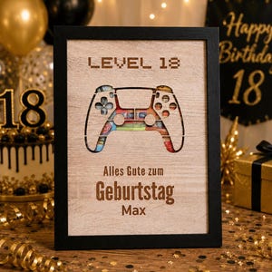 Könnte beinhalten: Ein rechteckiger Holzrahmen mit schwarzem Rand und einer hellen Holzplatte. Die Platte hat einen Ausschnitt in Form eines Videospiel-Controllers, in dem bunte Geldscheine sichtbar sind. Der Text lautet LEVEL 18, Alles Gute zum Geburtstag Max.