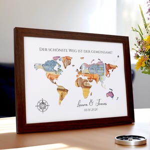 Könnte beinhalten: Ein gerahmter Kunstdruck einer Weltkarte mit einem dunkelbraunen Holzrahmen. Die Karte ist mit farbenfrohen Währungsdesigns gefüllt. Der Text "DER SCHÖNSTE WEG IST DER GEMEINSAME!" steht oben, darunter Namen und ein Datum.