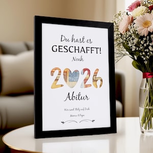Könnte beinhalten: Ein gerahmter Druck mit dem Text "Du hast es GESCHAFFT!" und "2026 Abitur". Die Zahlen sind mit bunten Mustern gefüllt. Der Druck befindet sich in einem schwarzen Rahmen und steht auf einer weißen Oberfläche. Ein Blumenstrauß ist im Hintergrund.