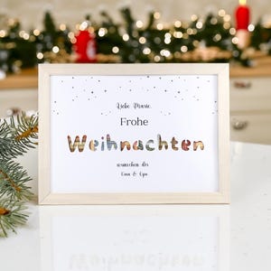 Könnte beinhalten: Ein gerahmter Druck mit dem Text "Liebe Marie. Frohe Weihnachten" in einer dekorativen Schriftart. Das Wort "Weihnachten" ist in bunten Buchstaben geschrieben. Der Rahmen ist aus hellem Holz und der Hintergrund ist unscharf.