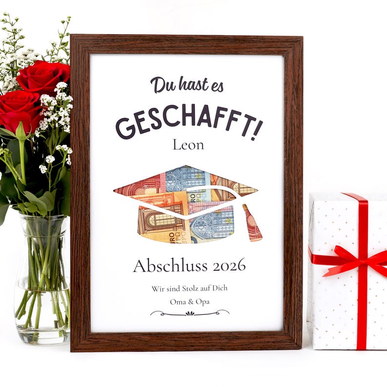 May include: A framed graduation print with the text "Du hast es GESCHAFFT!" and "Abschluss 2026". The artwork features a graduation cap made of Euro banknotes and a small bottle. A bouquet of red roses and a wrapped gift are also visible.