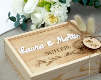 Op de afbeelding: Een lichtgekleurde houten kist met de namen "Laura & Martin" in witte letters en de datum "04.04.2026" gegraveerd. De kist is versierd met een bloemenontwerp en een klein houten label met de tekst "Alles Liebe".