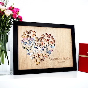Könnte beinhalten: Ein rechteckiges Wandbild aus Holz in einem schwarzen Rahmen mit einem herzförmigen Ausschnitt, der mit Schmetterlingssilhouetten gefüllt ist. Die helle Holzoberfläche ist mit dem Text Susanne & Niklas und dem Datum 26.06.2026 graviert.