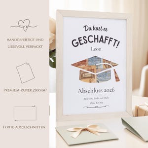May include: A framed graduation print with the words "Du hast es GESCHAFFT!" and "Abschluss 2026". The graduation cap is designed with currency. The print is in a light wood frame, with a light green card and bow in the foreground.