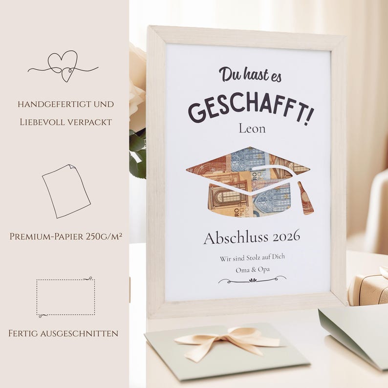 May include: A framed graduation print with the words "Du hast es GESCHAFFT!" and "Abschluss 2026". The graduation cap is designed with currency. The print is in a light wood frame, with a light green card and bow in the foreground.