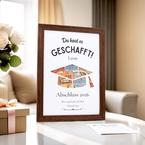 May include: A framed graduation print with a dark brown wooden frame. The print features the words "Du hast es GESCHAFFT!" and "Abschluss 2026". A graduation cap design is filled with colorful Euro banknotes. The print is a graduation gift.
