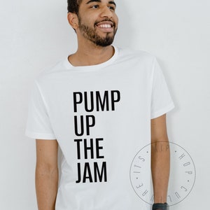 May include: White t-shirt with the text "PUMP UP THE JAM" in large, black capital letters. The shirt is a classic crew neck style. A circular graphic with text is visible on the lower right side of the shirt. The person wearing the shirt is smiling.