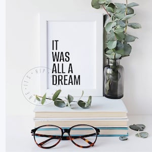 Puede incluir: Una impresi&oacute;n enmarcada en blanco con el texto "IT WAS ALL A DREAM" en letras negras. La impresi&oacute;n est&aacute; sobre una pila de libros con un par de gafas de montura marr&oacute;n delante de los libros. Una rama de vegetaci&oacute;n est&aacute; en un jarr&oacute;n de vidrio transparente a la derecha de la impresi&oacute;n.