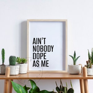 May include: A framed print with the text "Ain't Nobody Dope As Me" in black lettering on a white background. The print is displayed on a wooden shelf with potted plants.