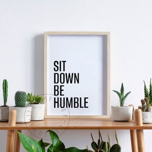 May include: A framed print with the text "SIT DOWN BE HUMBLE" in black lettering on a white background. The print is displayed on a wooden shelf with potted succulents.