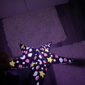 May include: A dark purple, five-pointed star-shaped paper lantern. The lantern is decorated with yellow stars, pink circles, white and blue shapes. The lantern is lit from within, casting a soft glow.
