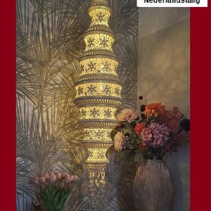 Può includere: Lampada a sospensione color crema con motivi floreali e design a più livelli. La lampada è illuminata, emettendo una luce calda. Lo sfondo presenta carta da parati floreale e un vaso di fiori. Il design della lampada è intricato.