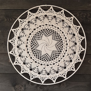 Op de afbeelding: Een witte gehaakte mandala met een spiraalvormig ontwerp in het midden, hangend aan een donker houten achtergrond.