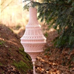 Könnte beinhalten: Ein rosa gehäkelter Lampenschirm mit einer Holzperle und einer Quaste, die an einem Baumzweig hängt.