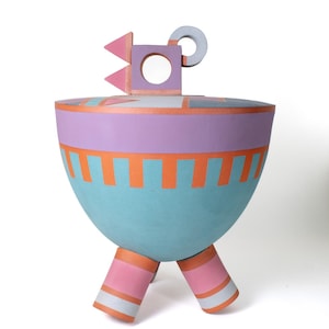 May include: A colorful, abstract ceramic vessel with a lid. The vessel is primarily light blue, with bands of lavender and orange. It has three pink legs and a gray handle. The lid features pink triangles and a circular opening.