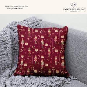 May include: A square decorative pillow with a festive pattern. The pillow features a deep red background with a repeating design of nutcrackers, gift boxes, and sprigs in shades of gold and cream. The pillow is displayed on a gray sofa with a knitted throw.